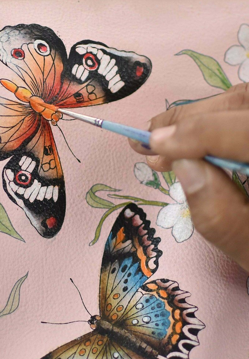 Schilderij van kleurrijke vlinders op een roze gestructureerd materiaal; fijne details omvatten levendige patronen en een hand die een kleine penseel vasthoudt.