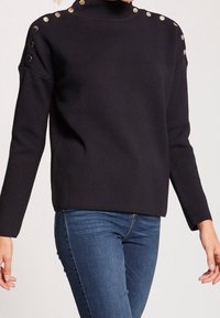 Zwarte gebreide trui met een hoge halslijn, met zilveren knoopaccenten op de schouders. Gestyled met blauwe jeans die een ontspannen pasvorm benadrukken.