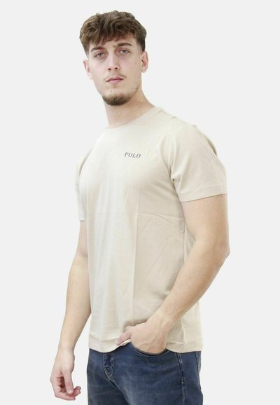Beige cotton t-shirt with short sleeves, crew neck, and a small "POLO" logo on the chest. Model wears blue jeans with a relaxed fit.
