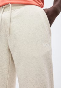 Κλειστό πλάνο μπεζ φόρμες με πλαϊνή τσέπη και κορδόνι, που φοριέται από άτομο με σκουρόχρωμο δέρμα και κοραλί πουκάμισο.