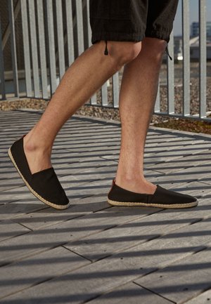 Person iført sorte slip-on sko og sorte shorts, der går på flisebelagt underlag med metalgelændere, der kaster stribede skygger.
