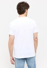 Tricou alb din bumbac cu un gât rotund, mâneci scurte și tiv drept, purtat cu blugi din denim de culoare albastră deschisă. Prezintă tatuaje vizibile pe braț.