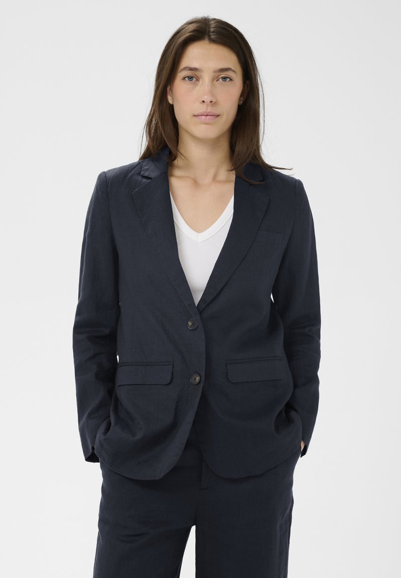 Moteris su tiesiais rudos spalvos plaukais, dėvinti tamsiai mėlyną švarką ir atitinkančias kelnes, po kuriais baltas V formos kaklo marškinėliai, stovinti su rankomis kišenėse.