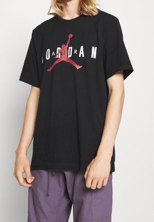 Person, die ein schwarzes Jordan-Brand-T-Shirt mit rotem Basketball-Silhouettenlogo und lila Hose trägt.