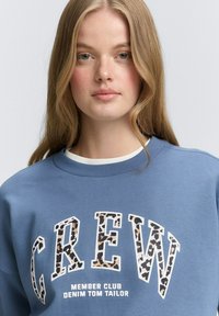 Felpa blu con scollatura a girocollo, caratterizzata da un logo "CREW" simile a una stampa leopardata e testo bianco sottostante. Texture morbida, vestibilità casual.