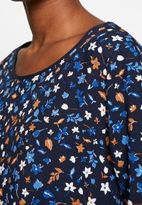 Top floreale con sfondo blu navy e piccoli fiori blu, bianchi e arancioni. Collo rotondo e texture in tessuto morbido.