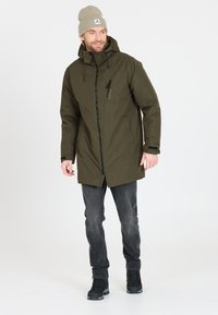 Olijfgroen waterdichte parka met een capuchon, voorrits en borstzak. Draag over grijze jeans en zwarte veterschoenen. Zachte beige beanie.