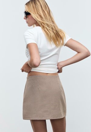 Donna bionda con occhiali da sole neri, t-shirt bianca aderente e mini gonna pied de poule beige, in piedi con le mani sui fianchi su uno sfondo neutro.