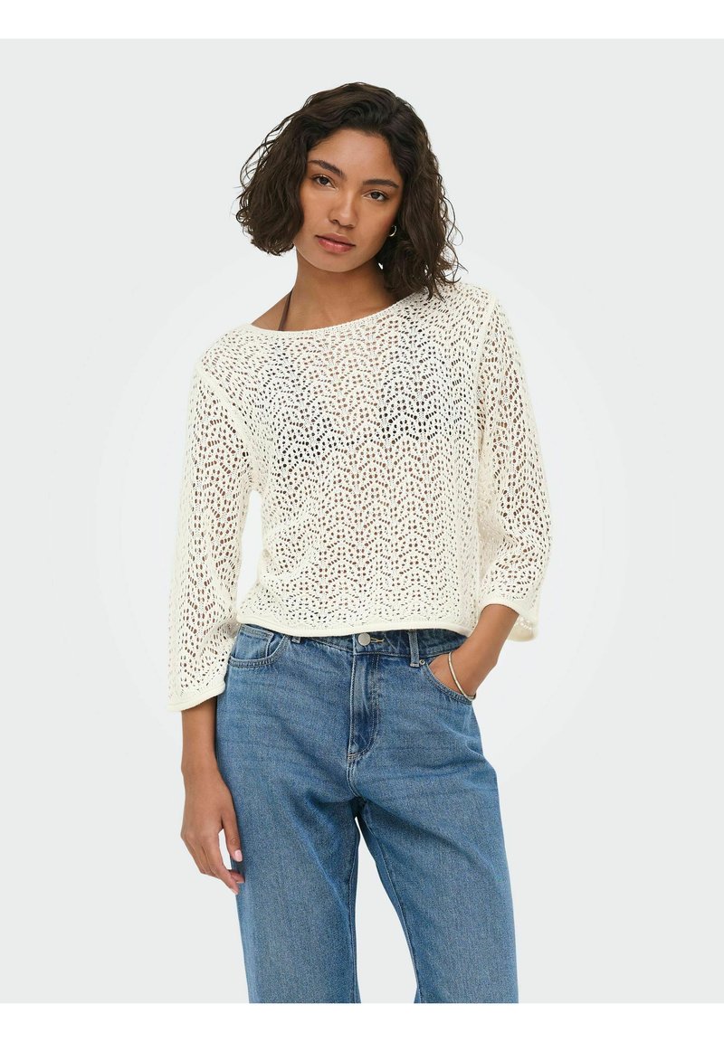 Femme aux cheveux courts et bouclés portant un haut blanc à manches longues en crochet et un jean bleu, debout avec une main dans la poche devant un fond uni.