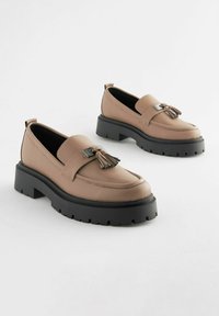 Mocasines de cuero beige con una suela de goma negra elevada, que presentan un borlas decorativas y un acento metálico en una correa ancha.