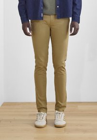 Schmal geschnittene khakifarbene Baumwollhosen mit glattem Material, kombiniert mit weißen Sneakers mit braunen Akzenten. Der Hintergrund besteht aus hellfarbigem Holz.