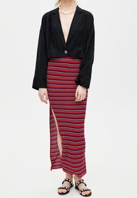 Černý zkrácený blazer s jedním knoflíkem, spárovaný s červenou pruhovanou maxi sukni s bočním rozparkem a černými sandály s kruhovými detaily.
