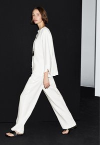 Vrouw met kort bruin haar die loopt, gekleed in een losse witte outfit en zwarte sandalen, voor een zwart-witte achtergrond.