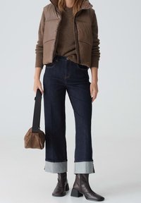 Kobieta w brązowym swetrze i kamizelce puchowej, w ciemnych spodniach z mankietami, ciemnych botkach za kostkę, trzymająca brązową torebk�ę z szerokim czarnym paskiem.