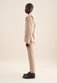 Completo in tessuto leggero color beige composto da una camicia con colletto e taschino sul petto, abbinato a pantaloni con risvolto e scarpe nere.