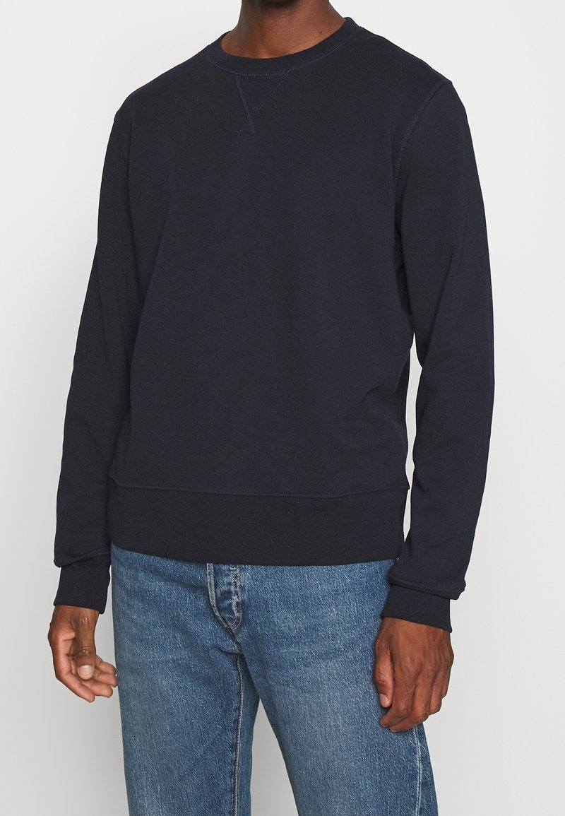 Homme portant un sweat-shirt à manches longues noir uni et un jean en denim bleu, debout devant un fond blanc.