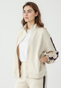 Beige Zip-Up-Jacke mit hohem Kragen, gerippten Bündchen und kontrastierenden dunklen Streifenakzenten an den Ärmeln. Weicher Stoff mit lässiger Passform.