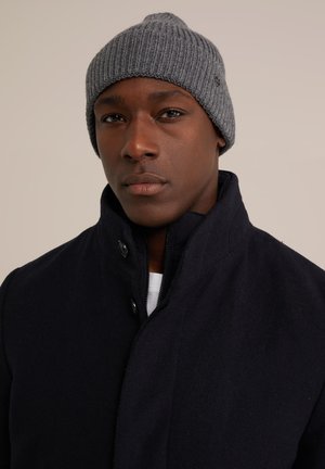 Black wool blend coat with high collar, featuring snap closures, layered over a white shirt. A gray ribbed knit beanie is worn on the head.