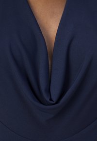 Marinblå klänning med djup V-ringning och draperad front, med slät tyg och subtil textur. Närbild visar detaljer vid halsringningen.