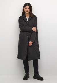 Cappotto imbottito lungo nero con colletto a ribalta, chiusura frontale con bottoni, tasche laterali e una texture liscia e elegante. Abbinato a pantaloni neri e stivali.