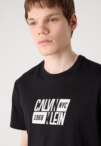 Fekete póló fehér grafikai dizájnnal, rajta a „CALVIN KLEIN NYC 1968” felirattal, vastag betűtípussal. Klasszikus kerek nyakkivágás és rövidujjú.