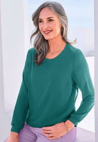 Langärmlige, smaragdgrüne Bluse mit rundem Ausschnitt und leicht durchscheinendem Stoff, kombiniert mit helllila Hosen und einem zarten Armband.