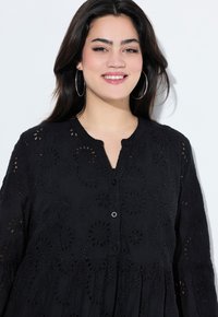 Mustat brodeeratut paidat, joissa on nappisuljenta, pyöreä kaula-aukko ja pitkät hihat. Kukkamaiset aukko-kuviot koko paidassa. Pehmeä rakenne.