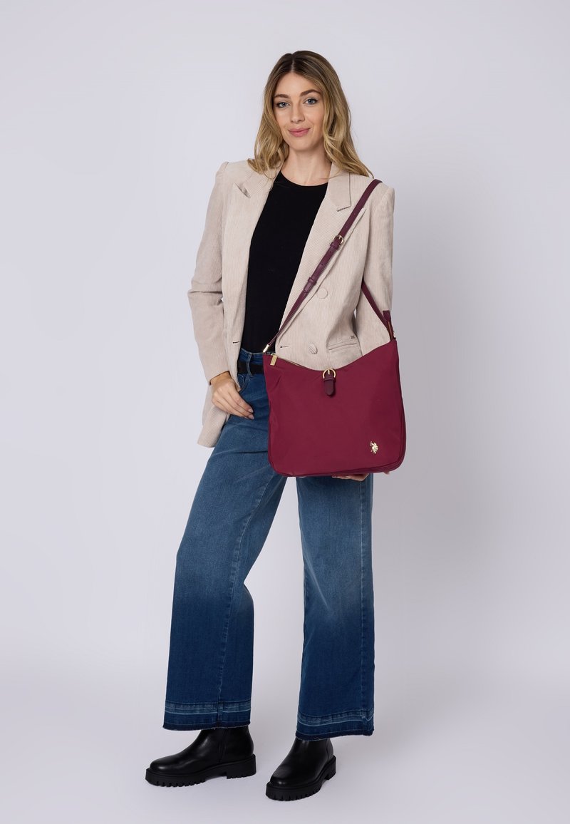 Borsa a spalla bordeaux con una texture liscia, dettagli in metallo dorato e una zip superiore. La borsa presenta un dettaglio con logo e una tracolla regolabile.