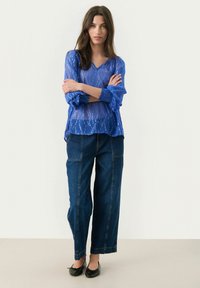 Durchsichtige blaue Bluse mit dezentem Muster, V-Ausschnitt und Puffärmeln, kombiniert mit locker sitzenden dunklen Jeanshosen. Schwarze Ballerinas runden das Outfit ab.
