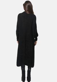 Lång svart stickad cardigan med lös passform, ribbad textur, vida ärmar och sidofrånskärningar; faller till midjan. Bärs över åtsittande byxor.