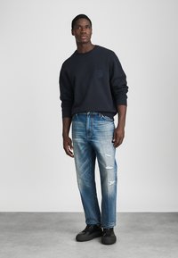 Ναυτικό φούτερ με λεπτομέρεια τσέπης, μπλε σκισμένα τζιν και μαύρα παπούτσια αθλητικά. Το ύφασμα φαίνεται μαλακό, με άνετη εφαρμογή και καθημερινό στυλ.