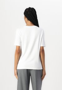 Vit kortärmad t-shirt gjord av mjukt tyg; har en rund halsringning och en avslappnad passform. Bärs tillsammans med grå skräddarsydda byxor.