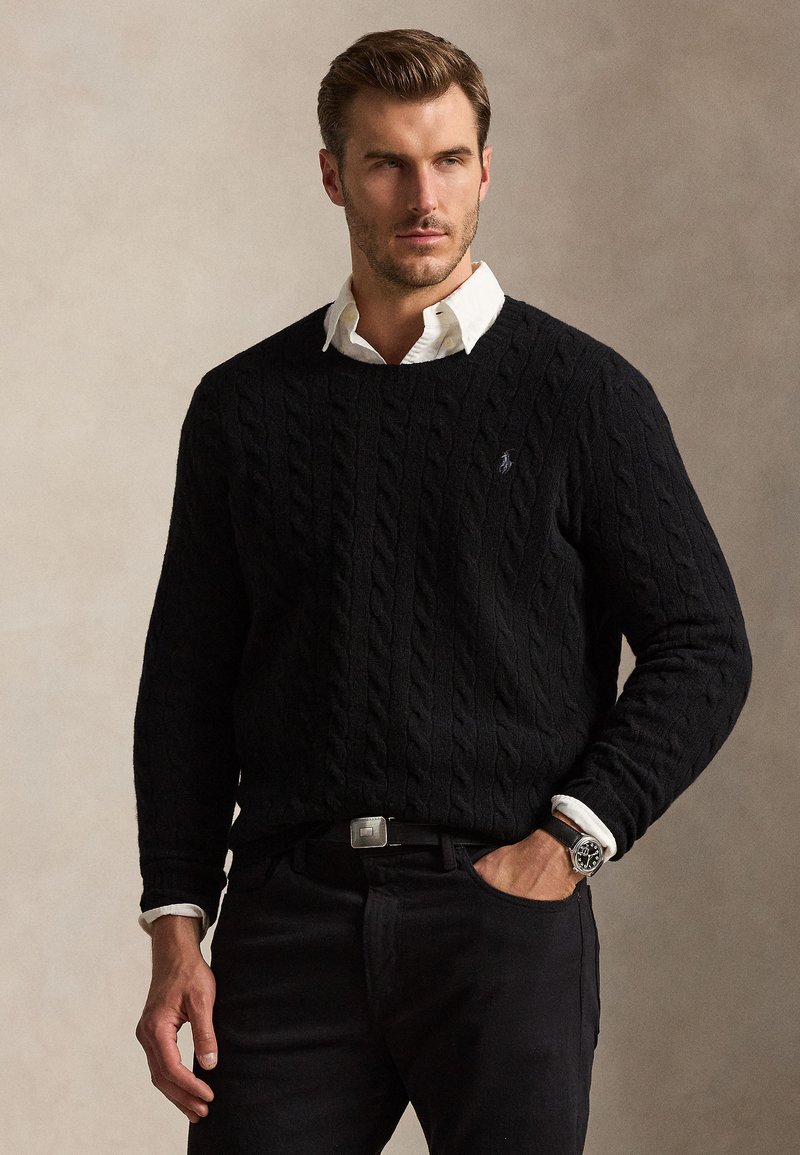 Uomo che indossa un maglione a trecce nero sopra una camicia bianca con colletto, pantaloni neri e orologio da polso, in piedi davanti a uno sfondo semplice.