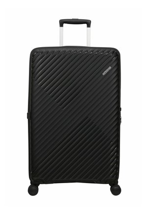 Grande valigia rigida nera con scanalature diagonali, quattro ruote girevoli, manico telescopico e logo "American Tourister" davanti.