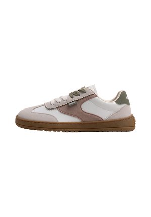 Zapatilla baja con paneles de ante blanco y beige, lengüeta verde en el talón, cordones bicolor y suela de goma marrón, mirando hacia la izquierda sobre un fondo blanco.