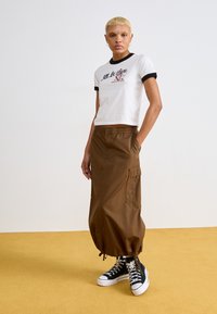 Witte cropped t-shirt met zwarte ringer hals en geborduurde tekst, gecombineerd met een bruine cargo rok met elastische taille en zwarte high-top sneakers.