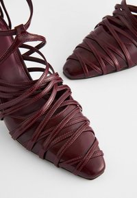 Burgundy strappy heels made of glossy leather with a pointed toe and crisscross strap design, featuring detailed stitching and a flat sole.