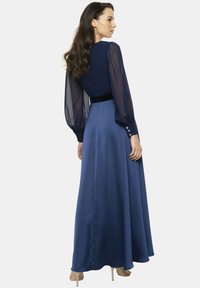 Długa niebieska sukienka z przezroczystymi długimi rękawami i aksamitnym paskiem w talii. Gładki satynowy materiał o zwiewnym kroju. Okrągły dekolt i akcenty w postaci guzików.