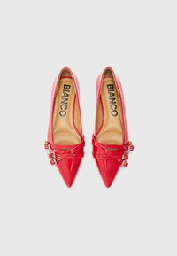 Chaussures à talons aiguilles rouges avec des orteils pointus et des sangles croisées avec des boucles, présentées sur un fond blanc uni.