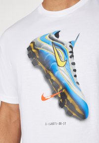 T-shirt bianca con una grafica di una scarpina da calcio blu e gialla, con dettagli neri e un logo Nike arancione sotto la scarpa.