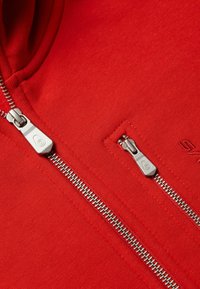 Červená mikina s hladkým materiálem, která má vpředu dva stříbrné zipy, jeden je částečně rozepnutý, a detail kapsy se švem.