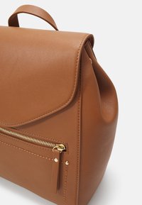 Sac à dos en cuir marron avec rabat supérieur, poche zippée avant avec fermeture éclair dorée, et poignée de transport supérieure sur fond blanc.