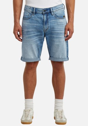 Man draagt lichtblauwe opgerolde spijkershorts, witte sokken en beige sneakers, staand tegen een effen achtergrond.