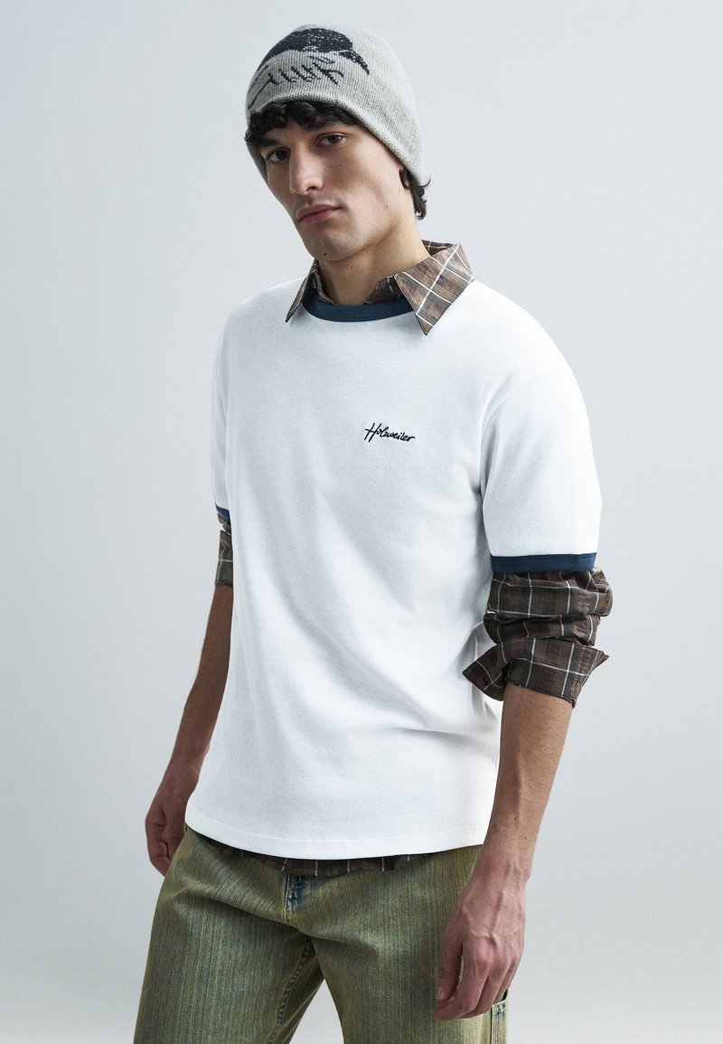 Fehér rövid ujjú póló tengerészkék részletekkel és hímzett logóval, barna kockás flanel ingre rétegezve, világoszöld farmernadrággal párosítva.