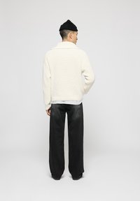 Cárdigan de punto crema con un cuello ancho, combinado con jeans oscuros de corte holgado. La modelo lleva un gorro negro, de pie frente a un fondo neutro.