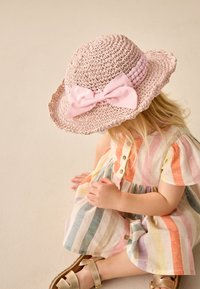Chapeau de paille rose pâle avec un large bord et un bord festonné, orné d'un grand nœud. Enfant portant une robe rayée multicolore.