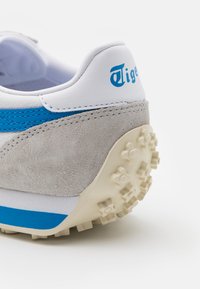 Sportovní tenisky s bílým koženým svrškem, modrými akcenty, šedými semišovými detaily a texturovanou béžovou gumovou podrážkou s gripovými vzory.
