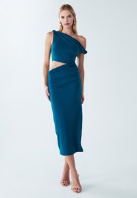 Obleka v barvi modrozelene, z eno ramo, iz gladkega blaga, s poudarki izrezov in naborkov. Elegantna, dolga do tal z oprijetim krojem.