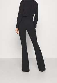 Schwarze, hochtaillierte, ausgestellte Pants mit glatter Textur, Nahtdetails und weitem Beinöffnungsverlauf. Die Rückansicht zeigt keine sichtbaren Taschen.