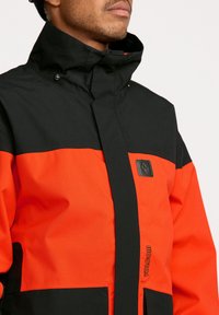 Veste résistante à l'eau avec un design en blocs de couleurs noir et orange, col haut, boutons-pression et patch logo sur la poitrine.